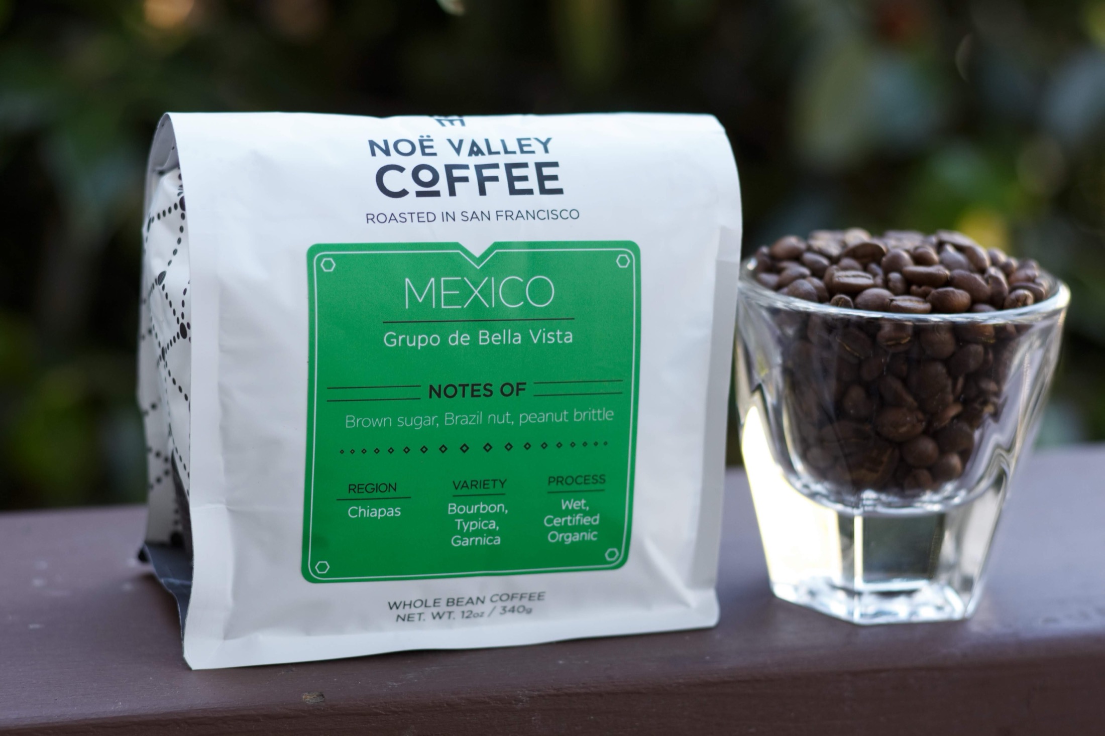 A 12 ounce bag of Noe Valley Coffee Mexico Chiapas beside a glass of roasted coffee beans.